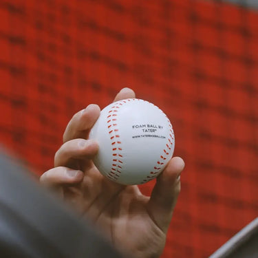 Hand holding white foam baseball with red stitching, labeled 'FOAM BALL BY TATER®' against red net background