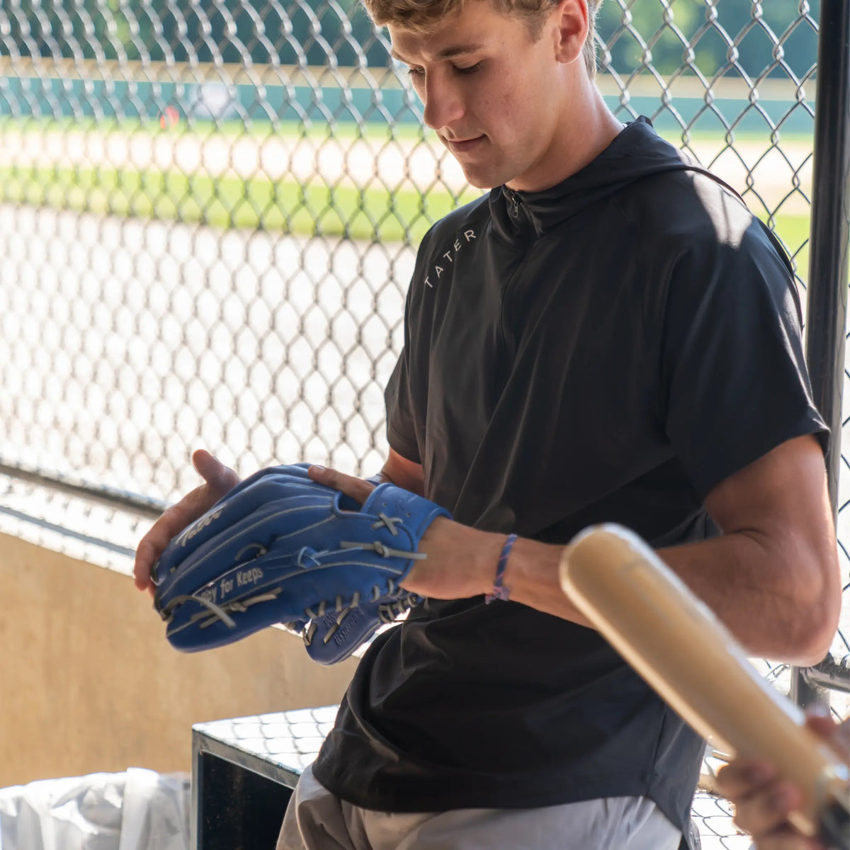 A baseball player is seen wearing a black short-sleeve BP (batting practice) hoodie from Tater Baseball. He is holding a blue outfielder&#39;s glove and a natural wood bat, indicating he&#39;s possibly preparing for practice or a game.