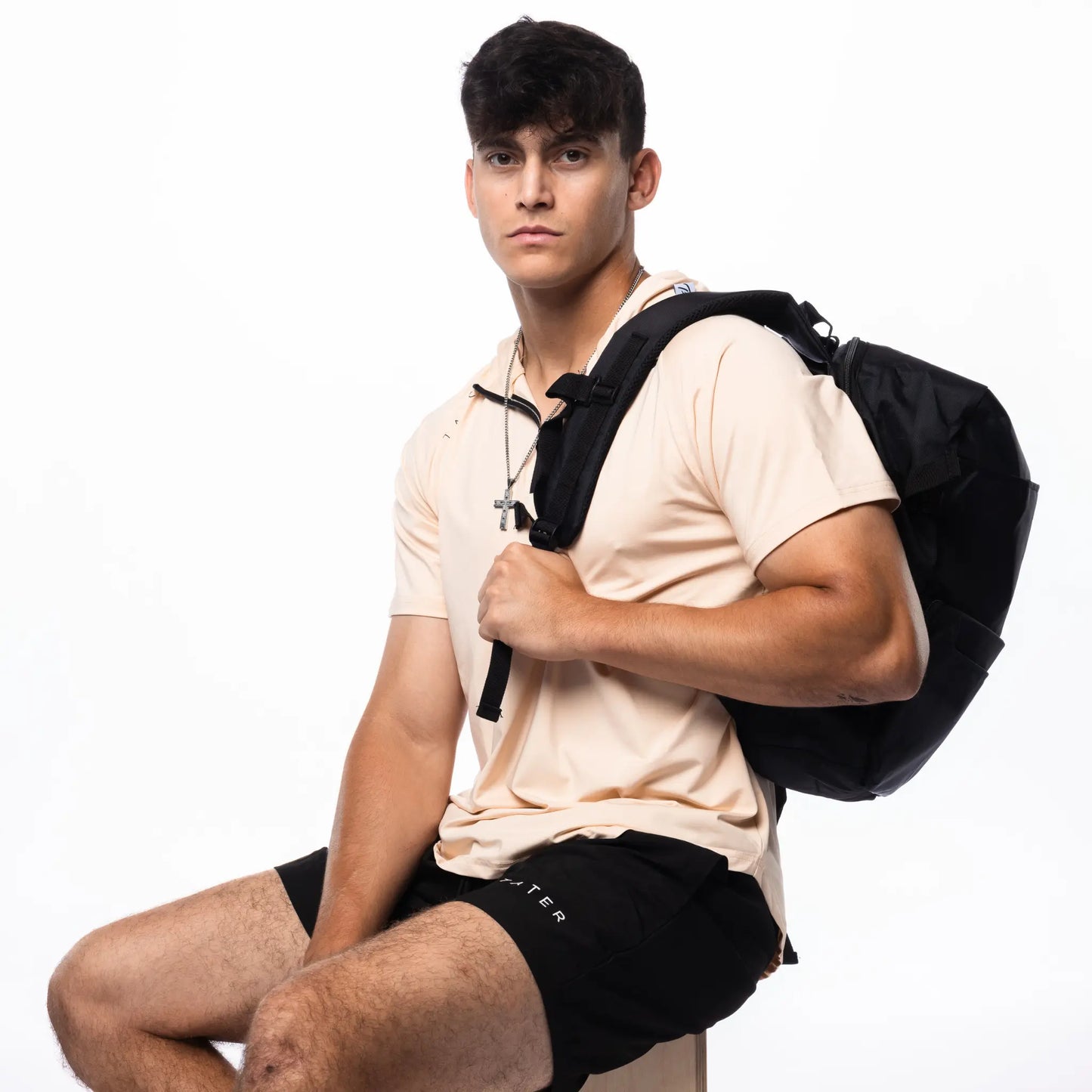 In the image, there is a young man wearing a tan short-sleeved hoodie and black athletic shorts, both featuring the "TATER" logo, which suggests they are part of a baseball athletic wear line. He is also carrying a black backpack over one shoulder and has a set of earphones around his neck. His attire and accessories indicate a casual, sporty aesthetic, likely targeting athletes or individuals with an active lifestyle.