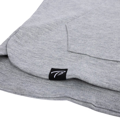 Detail shot of the hem on a Tater Baseball grey long-sleeve hoodie, featuring the brand's distinctive TB logo on a small black tag, highlighting the brand's subtle yet stylish detailing.