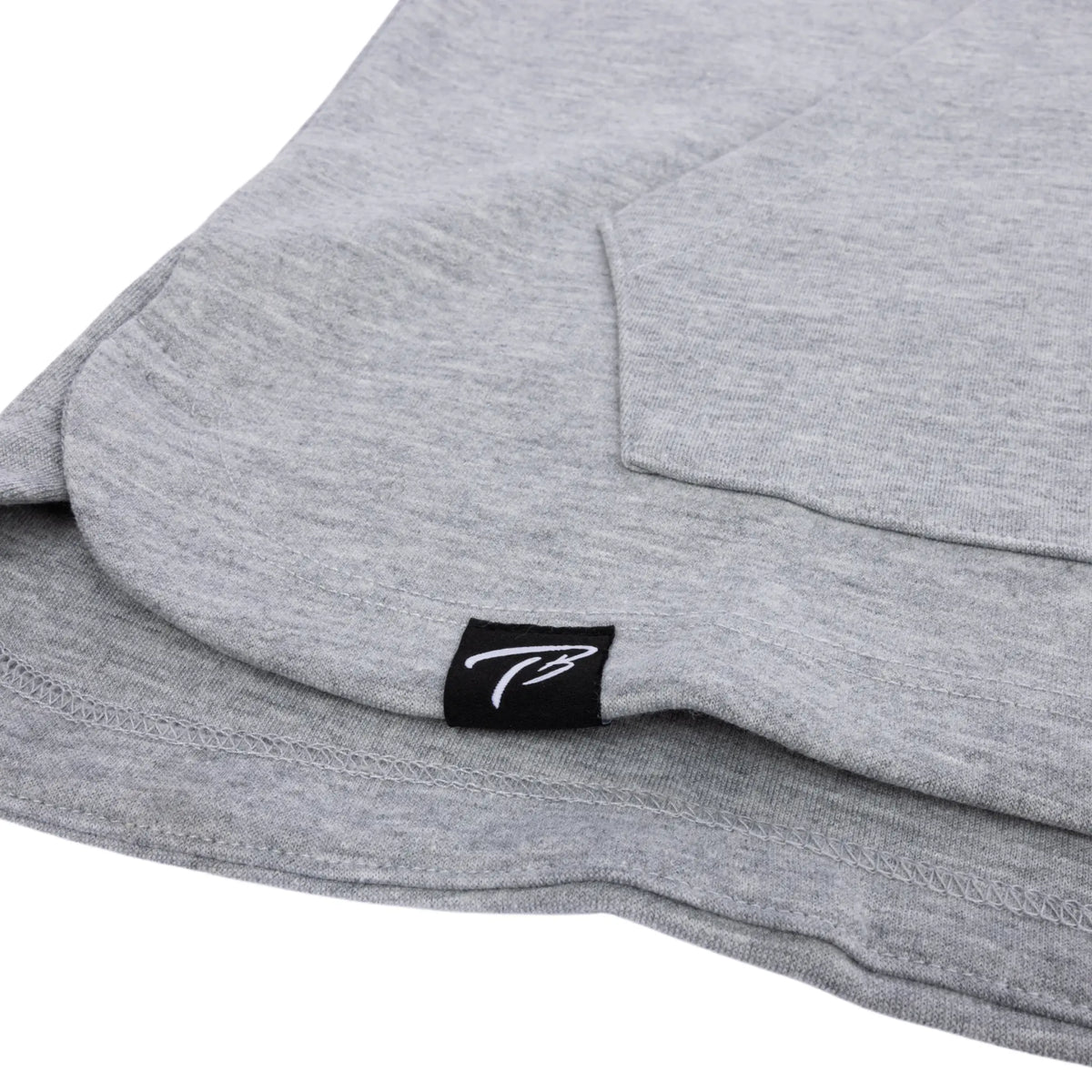 Detail shot of the hem on a Tater Baseball grey long-sleeve hoodie, featuring the brand&#39;s distinctive TB logo on a small black tag, highlighting the brand&#39;s subtle yet stylish detailing.