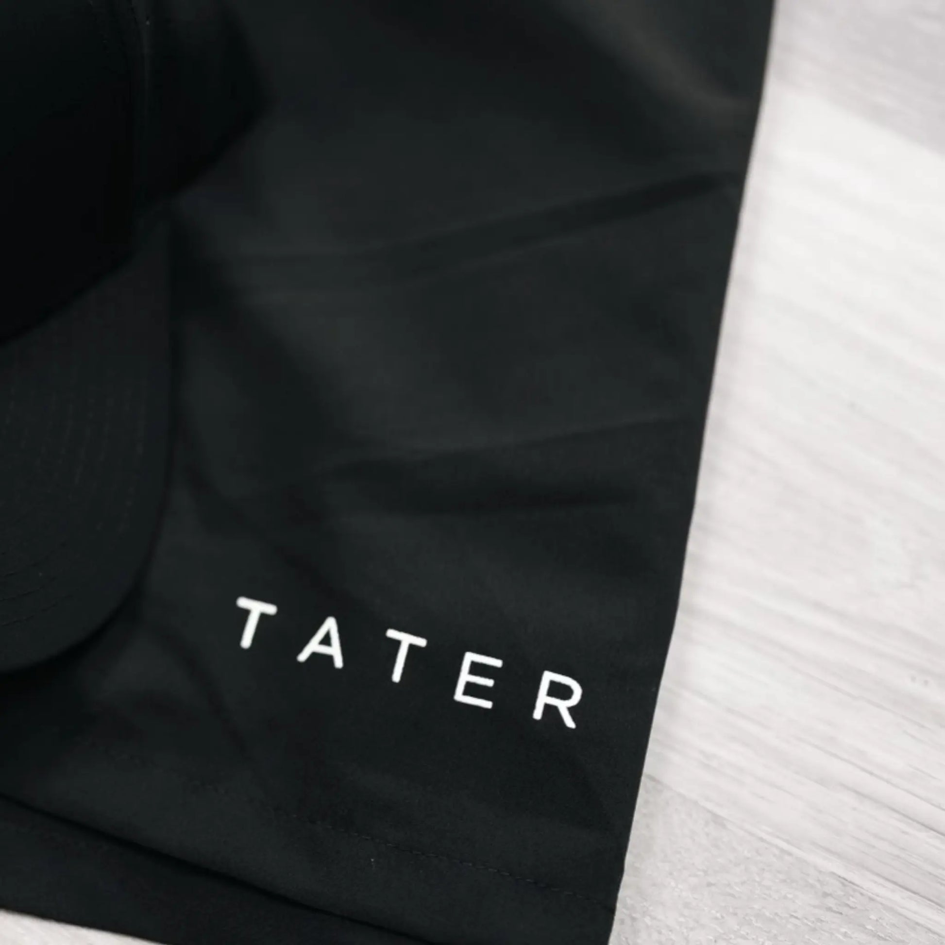  Image shows a close-up of black athletic shorts with the brand name "TATER" printed in white letters, likely indicating a focus on sports or fitness apparel. The material and design suggest that these shorts are made for active use, suitable for workouts, baseball practice, or casual wear. The branding is simple and elegant, which could appeal to consumers looking for stylish yet functional athletic wear.