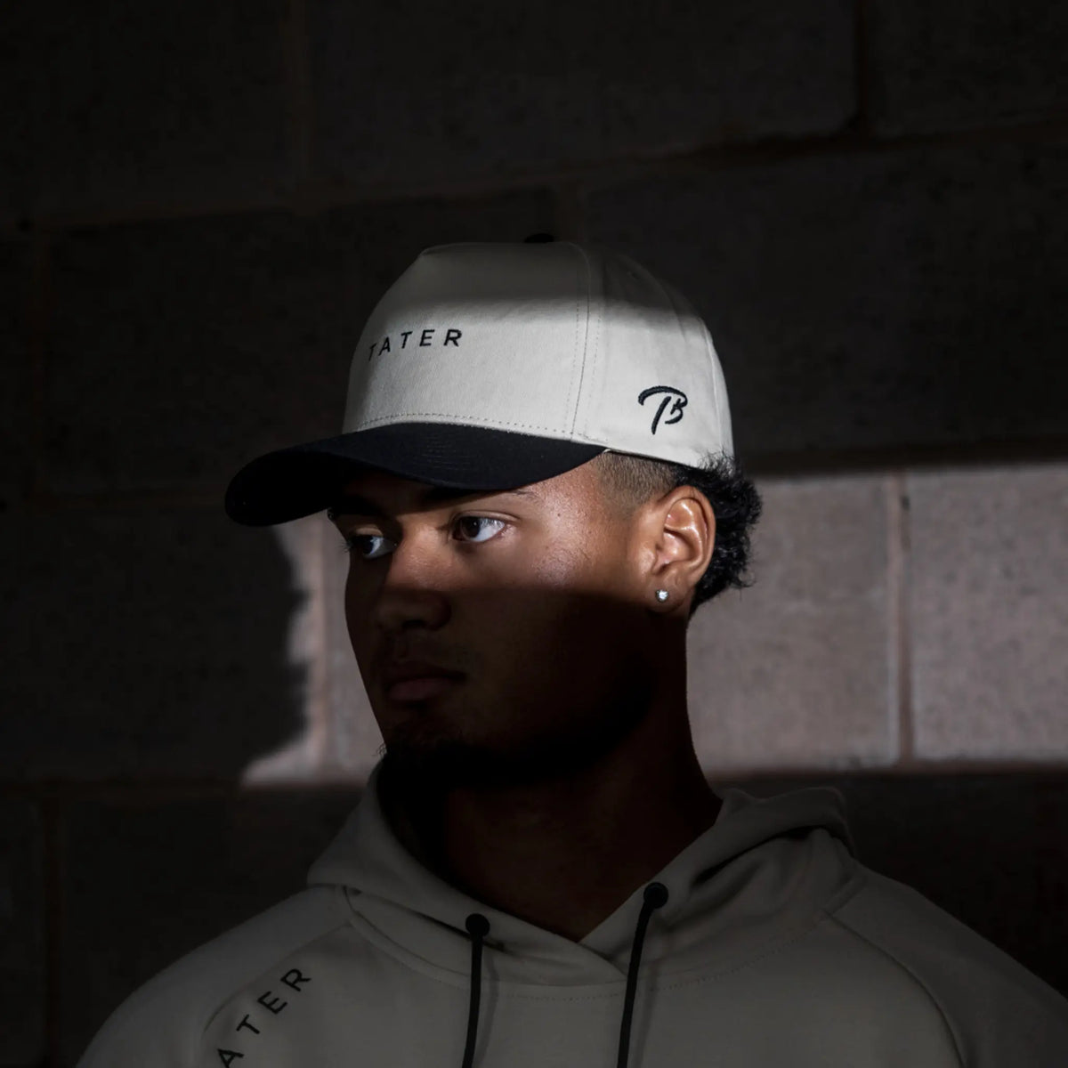 The image features a person wearing a tan snapback hat with the word &quot;TATER&quot; on the front and the Tater Baseball &quot;TB&quot; logo on the side, paired with a hoodie that also carries the &quot;TATER&quot; branding. The overall look suggests a coordinated athletic style, with the person looking off to the side, creating a contemplative and focused atmosphere. The lighting in the photo highlights the hat and the person&#39;s profile, adding to the moody and professional aesthetic of the apparel.