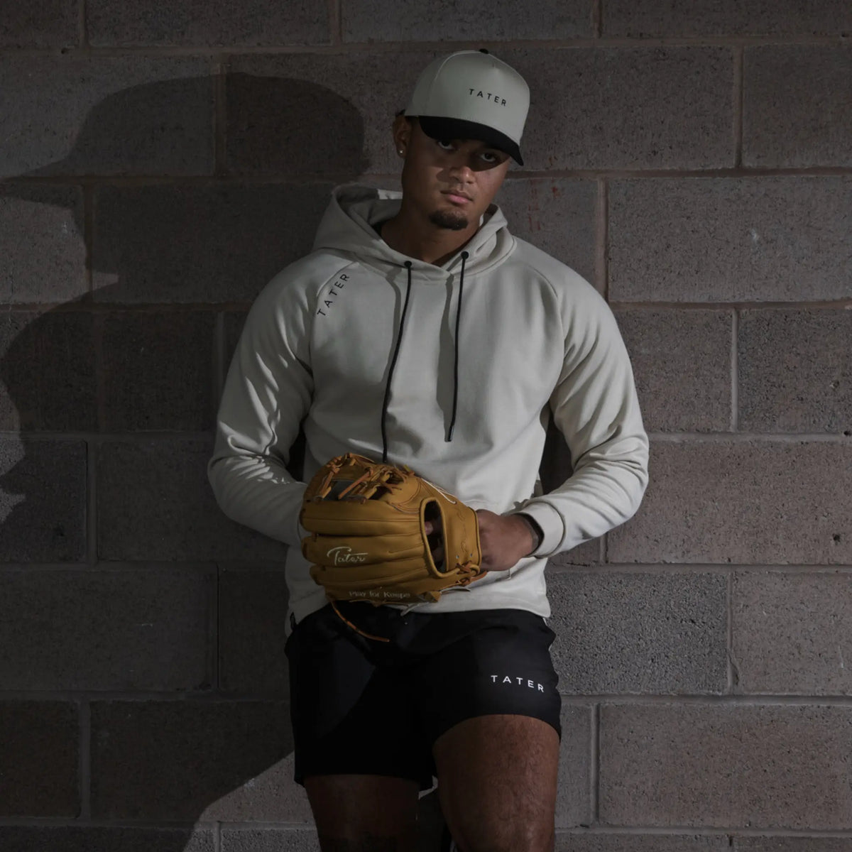 The image showcases an individual donning a coordinated sports ensemble from Tater Baseball. The person is wearing a cream snapback hat with the brand&#39;s name and logo, complemented by a matching hoodie. In one hand, they hold a gold-colored baseball glove, and they&#39;re dressed in black athletic shorts bearing the brand&#39;s name. The overall styling presents a sleek, athletic look, with a backdrop that adds a raw, urban feel to the image, highlighting the sporty yet stylish nature of the apparel. 