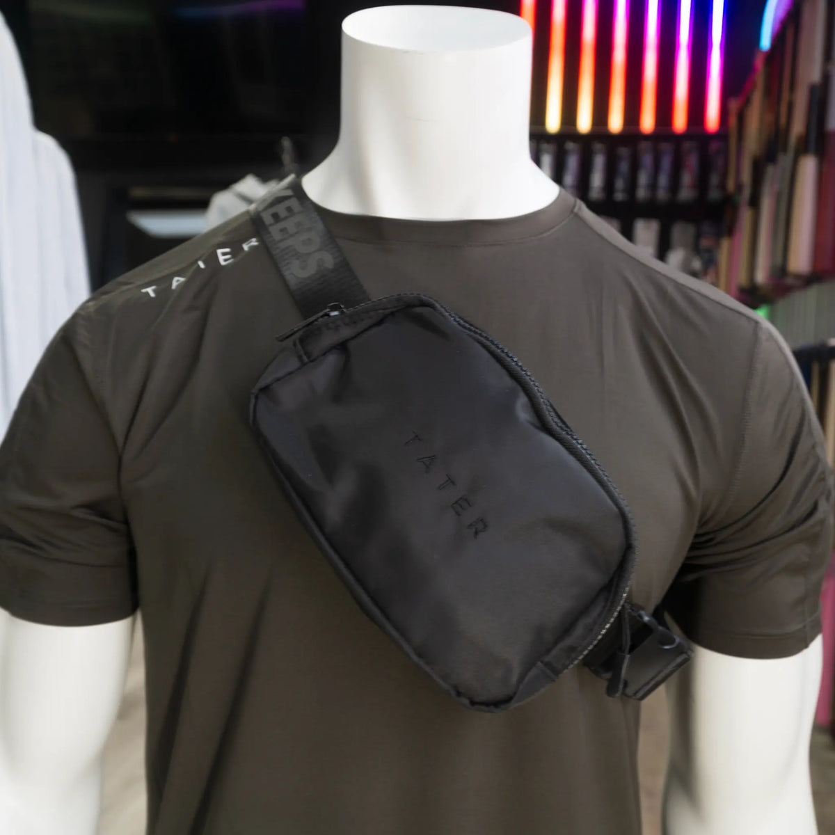 The image presents a mannequin dressed in an olive athletic T-shirt with a black crossbody fanny pack slung over the shoulder. The fanny pack features the Tater brand name embossed on the front, while the strap is adorned with the phrase &quot;KEEPERS&quot; in a repeating pattern, contributing to the sporty and stylish aesthetic of the product. The combination of the crossbody pack with the athletic wear suggests a trendy and practical accessory for sports enthusiasts or individuals with an active lifestyle. 