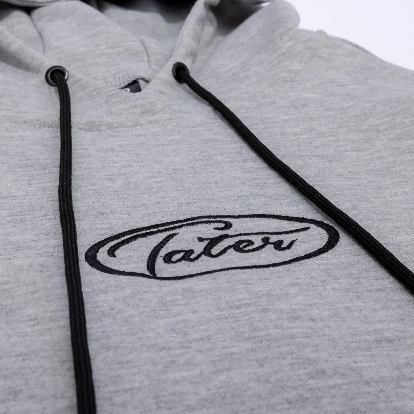 Close-up of a grey Tater Baseball hoodie featuring the brand's oval logo in black, complemented by contrasting black drawstrings, highlighting the brand's chic, sporty aesthetic