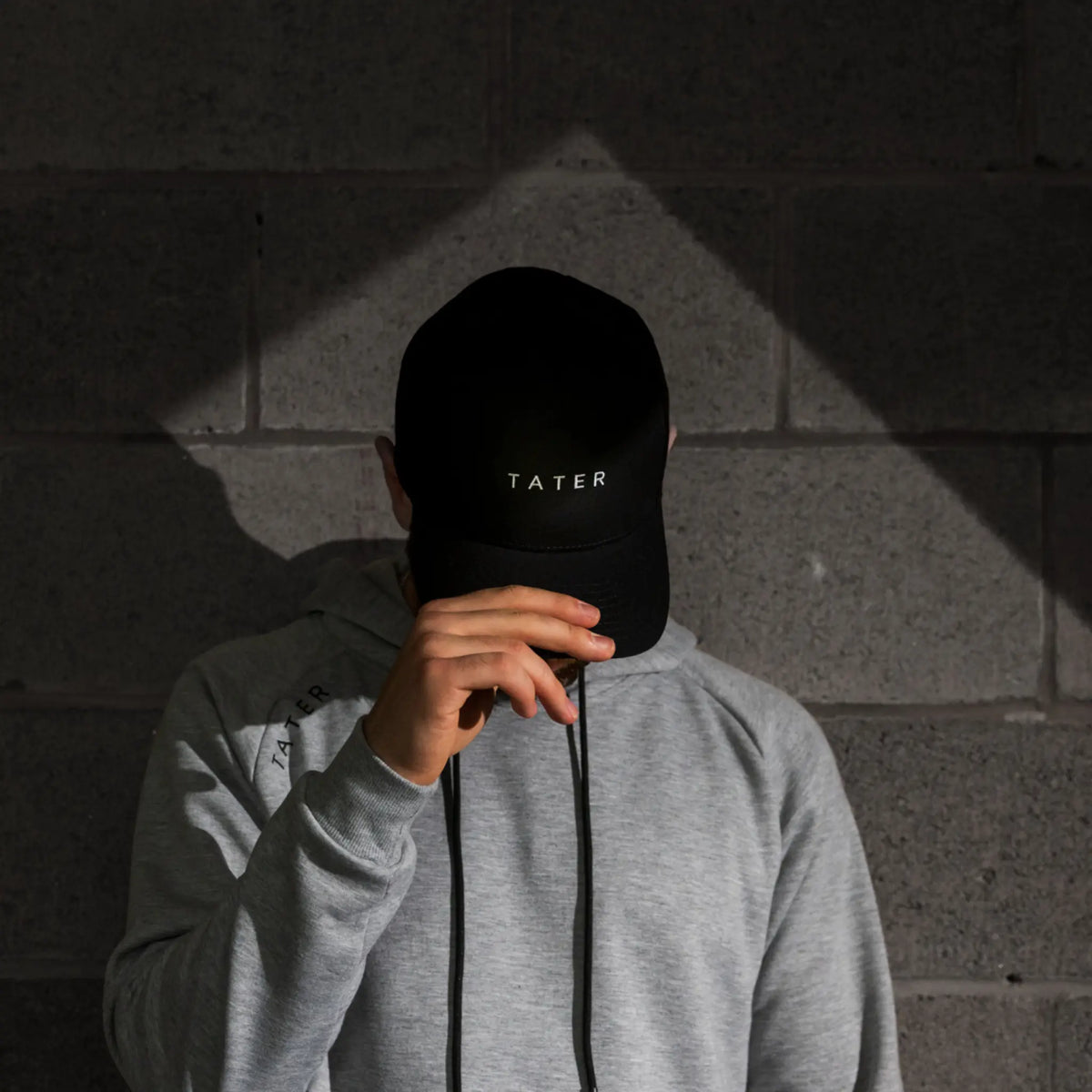 This image presents a person in a shadowy environment wearing a grey long-sleeve shirt and holding a black snapback cap with &quot;TATER&quot; embroidered in white across the front. The style is modern and minimalist, appealing to those who appreciate understated, sleek fashion. The cap is part of the Tater Baseball lifestyle collection, indicating a blend of sports and casual wear. The lighting and setting give the image an edgy, stylish vibe.
