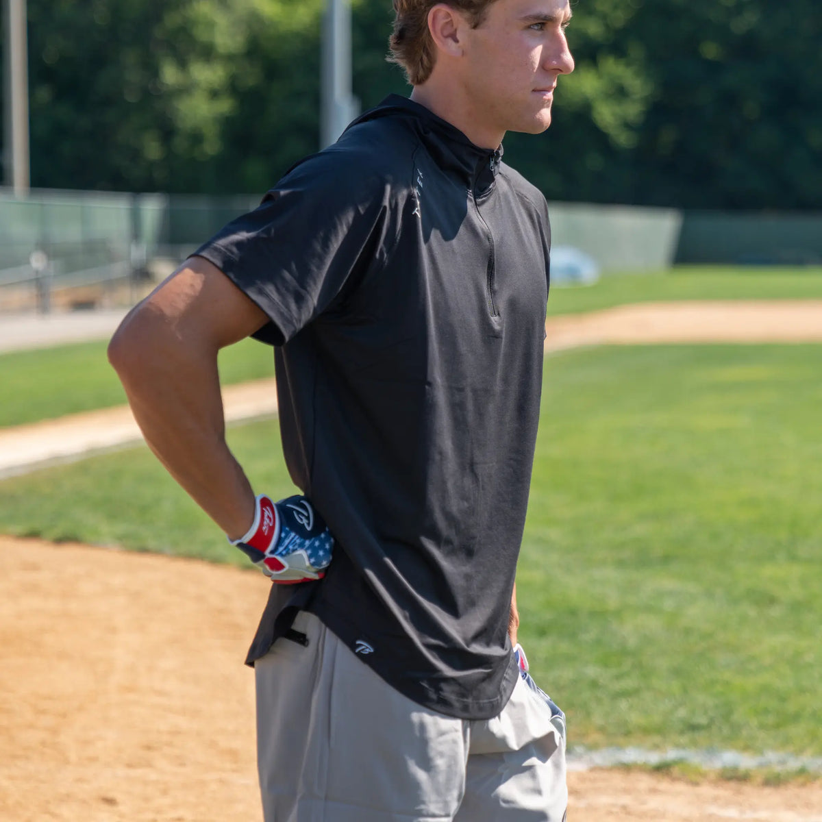 Baseball player in a reflective pose wearing a black short-sleeve hoodie and white baseball pants, equipped with a batting glove, contemplating his next turn at bat on a sunny field.