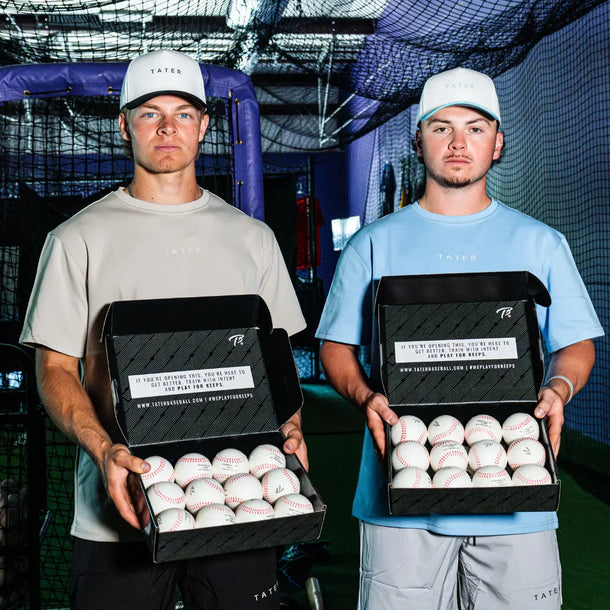 Two players holding open boxes of Tater foam pitching machine balls, two dozen per box, in an indoor batting cage